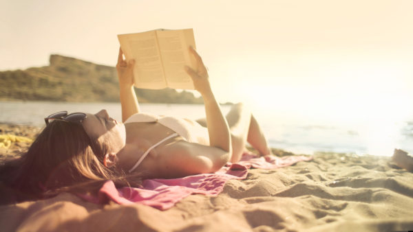 Sahilde bir kız uzanmış ve güneş ışığının altında denize karşı kitap okuyor