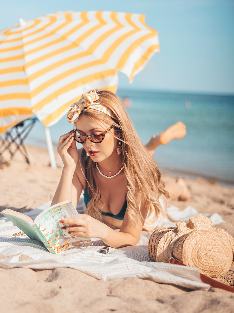 sahilde güneş gözlükleriyle uzanıp güneşlenirken kitabını okuyan kızın resmi