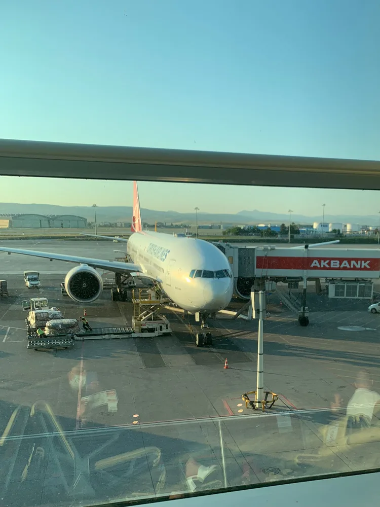 Camın arkasında kalkışa hazırlanan bir uçak ve saatin dolmasını bekleyen yolcu
