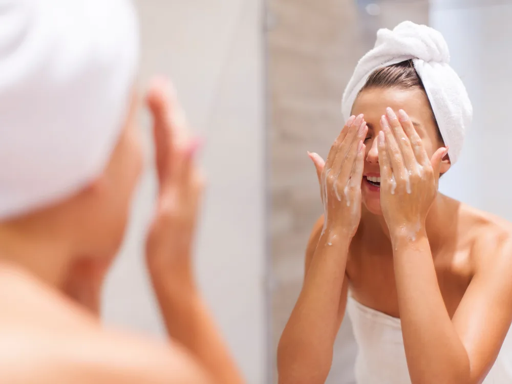 Aynanın karşısında yüzünü yıkayarak cilt bakımı yapan beyaz havlulu kadın,